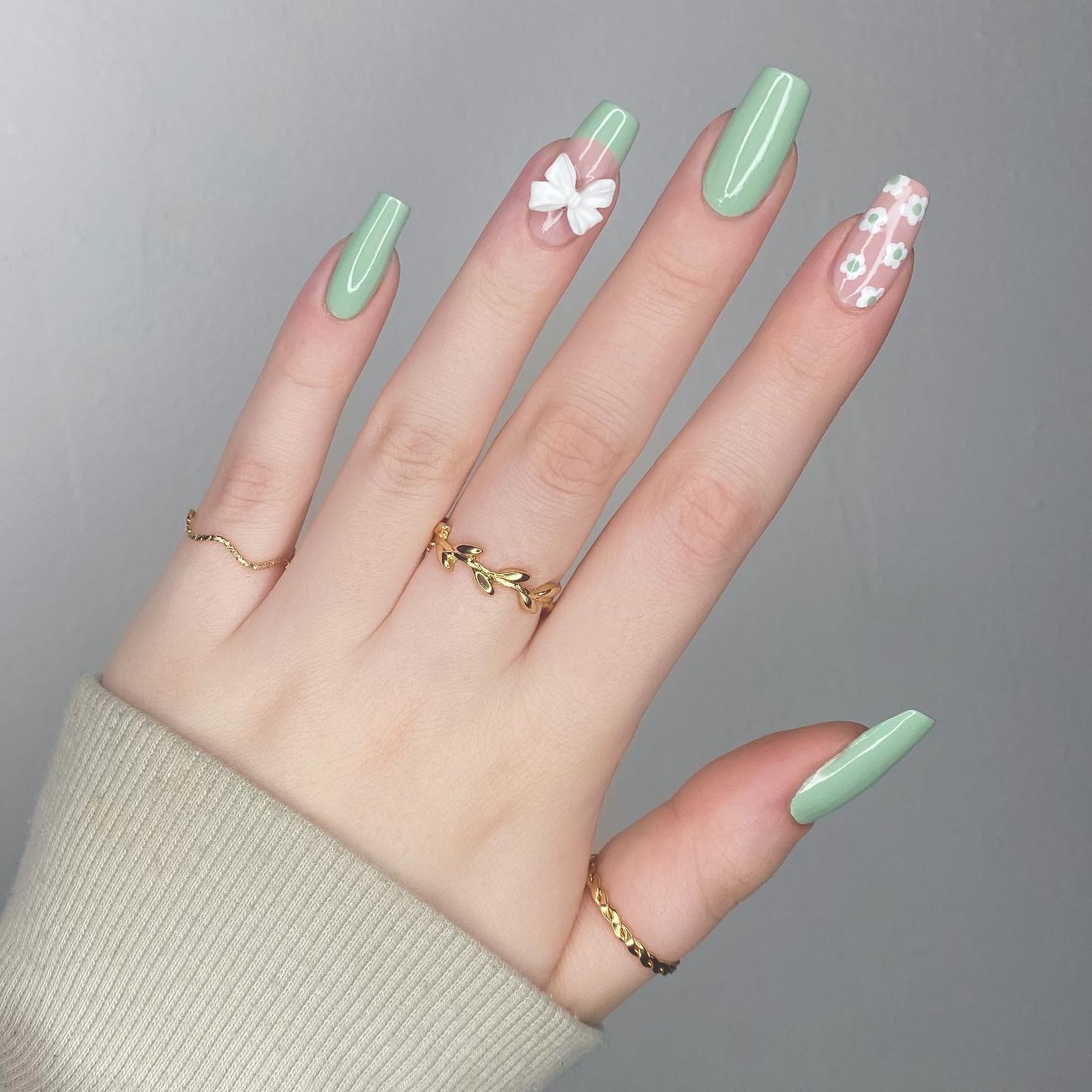 Eine Nahaufnahme salbeigrüner Nägel mit French-Tip-Akzent, weißen Blumen und Applikationen