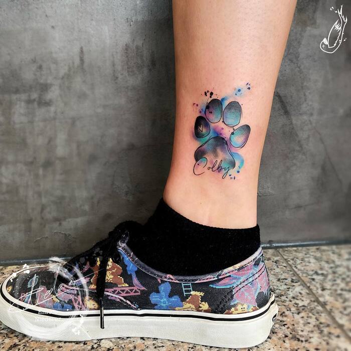 Tattoo of black dog’s print with blue and purple watercolor splashes and dog name under tattoo
