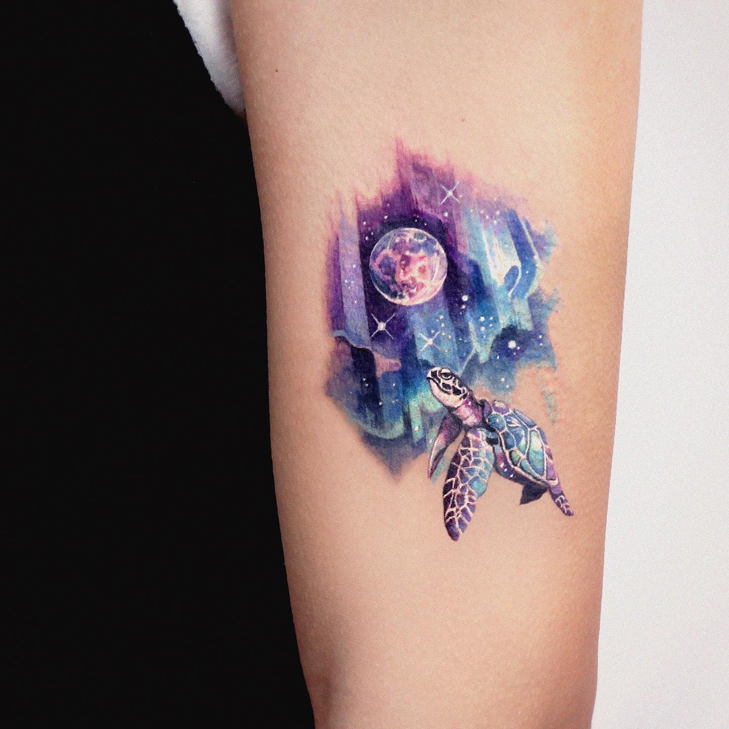 Akwarelowy tatuaż przedstawiający tło w formie przestrzeni z planetą, do której dryfuje żółw
