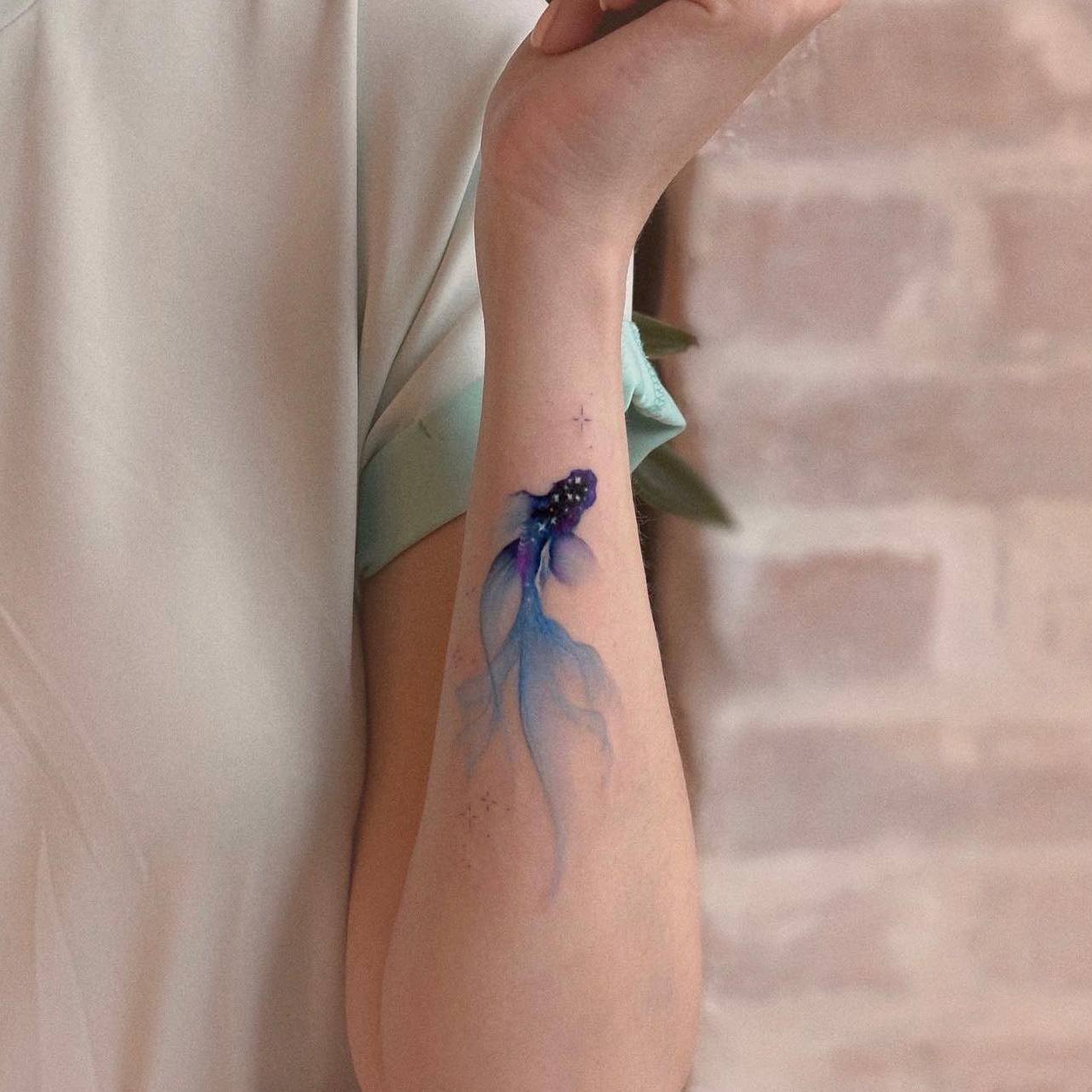 Akwarelowy tatuaż przedstawiający rybę koi na nadgarstku, wykonany tuszami w kolorze głębokiego fioletu i błękitu