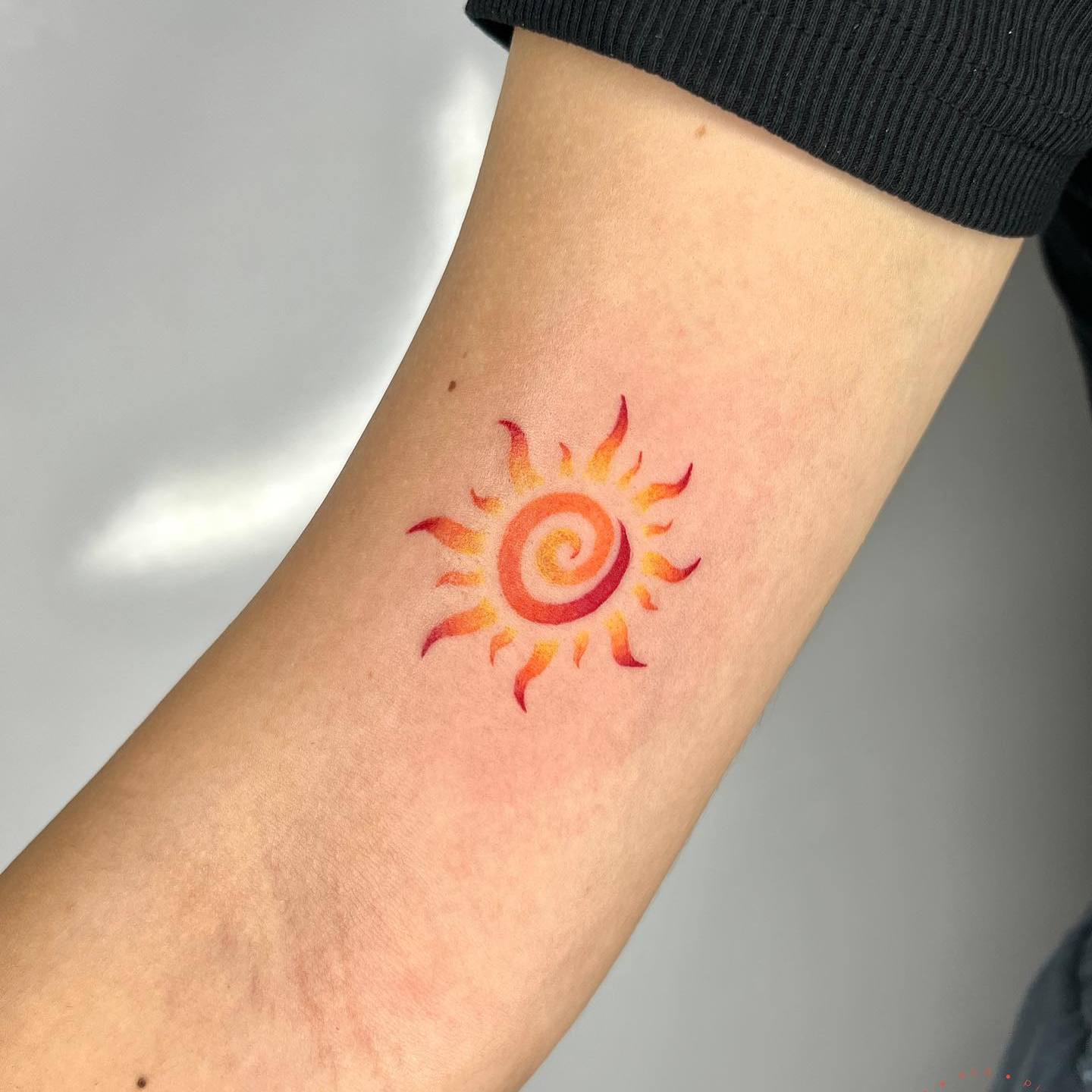 Akwarelowy tatuaż przedstawiający małe pogańskie słońce, wykonany tuszami w kolorze czerwonym, pomarańczowym i żółtym