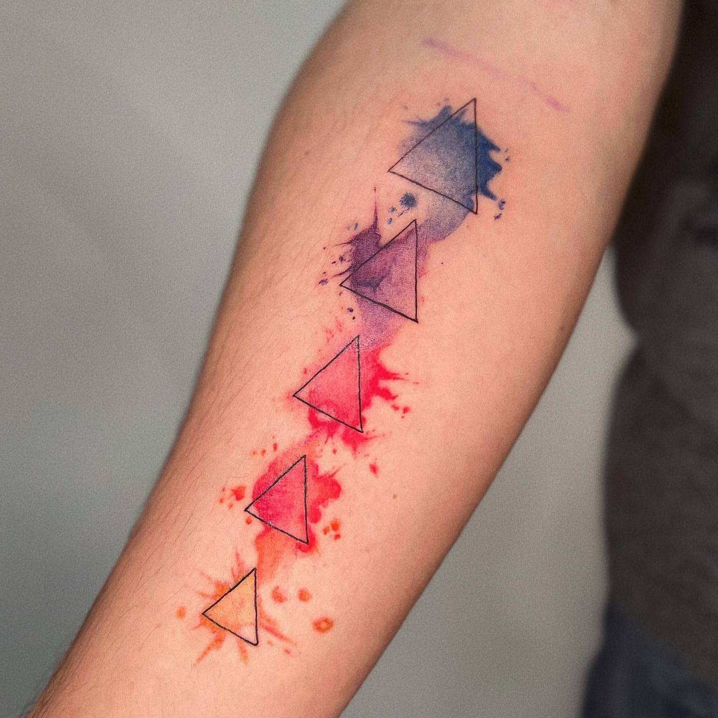 Akwarelowy tatuaż przedstawiający pięć trójkątów o różnych rozmiarach i kolorach, ułożonych jeden za drugim
