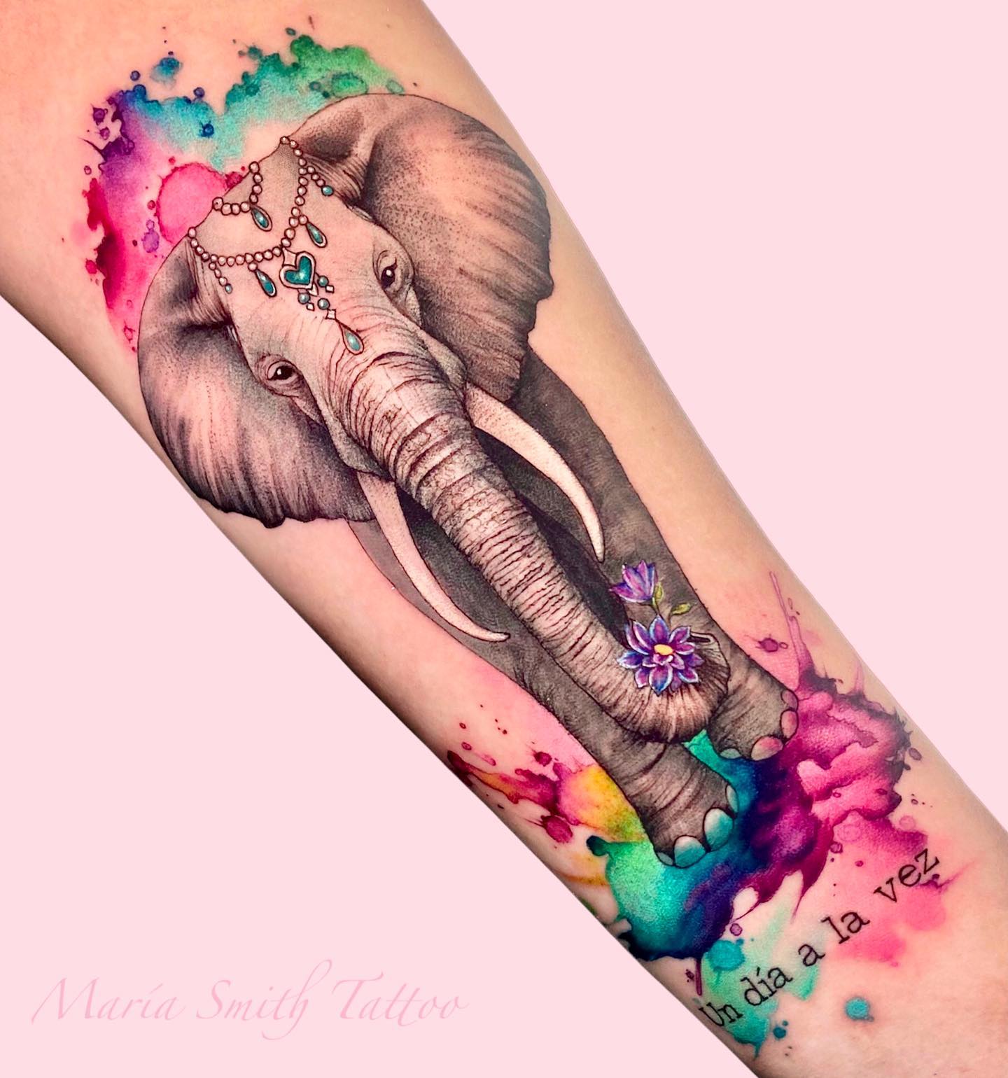 Akwarelowy tatuaż przedstawiający szarego słonia niosącego kwiaty z jasnymi kolorowymi plamami dookoła
