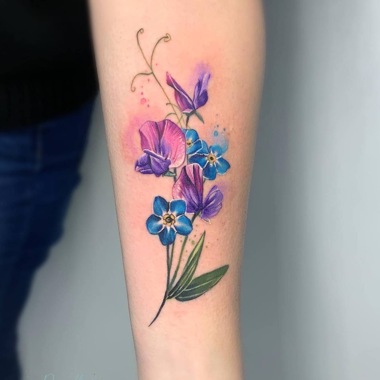 Akwarelowy tatuaż przedstawiający fioletowe i niebieskie kwiaty z zieloną rufą