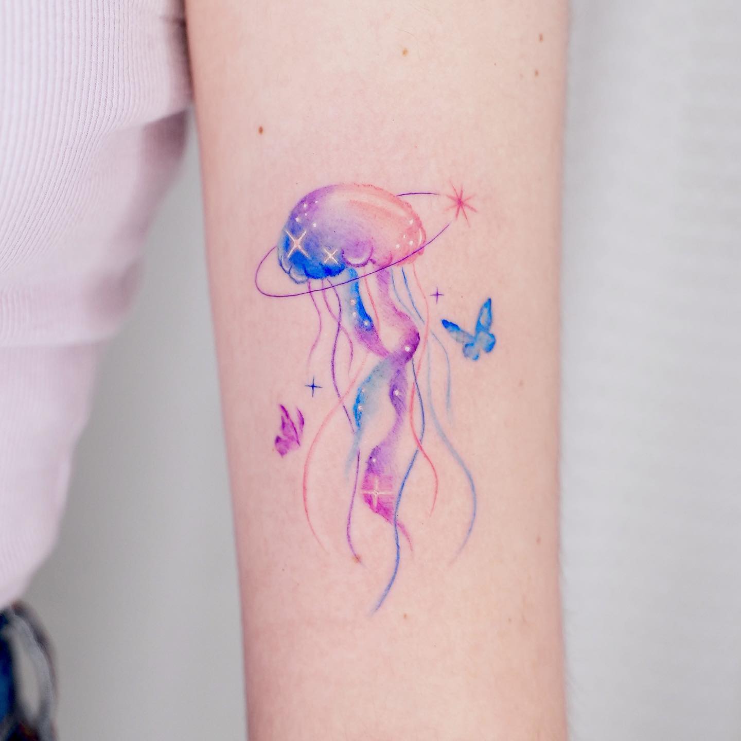 Akwarelowy tatuaż z wypełnioną niebieską, fioletową i pomarańczową meduzą oraz dwoma małymi motylami